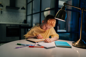 Front view of focused pupil student boy studying at home reading paper book, doing homework, learning sitting at table under light of lamp at night. Schoolboy reading textbook sitting at desk.