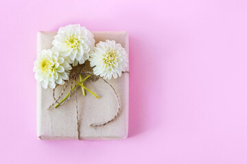 Gift box in gray craft paper decorated with white dahlia flowers on a pink background for the holiday.