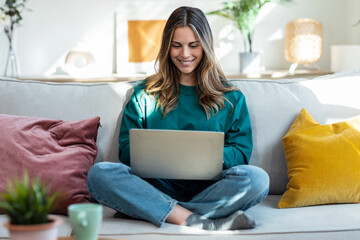 Beautiful kind woman working with laptop while sitting on couch in living room at home - obrazy, fototapety, plakaty