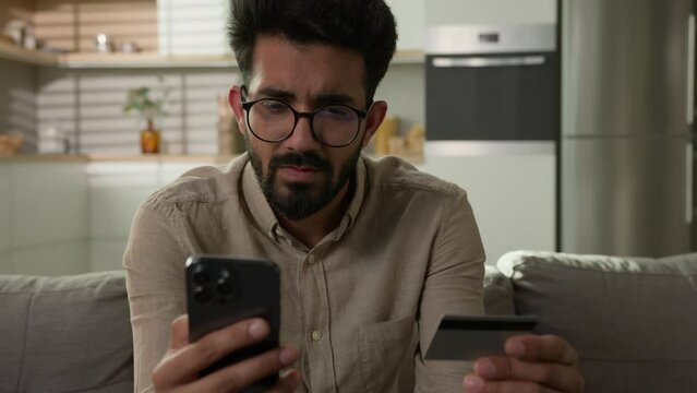 Arabian Indian man on sofa confused upset business client at home kitchen problem online payment credit limit use mobile phone app and bank card failure mistake error trouble not enough money balance