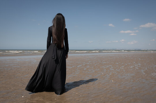 fine art rear view of woman in classic black dress on beach near ocean