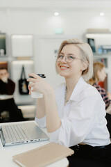 Young beautiful joyful blonde woman smiling while working with laptop in cafe with coffee, white light