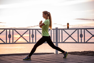 Young woman doing lunges, squats on warm-up in morning at sunrise by sea. Sportswoman in sportswear