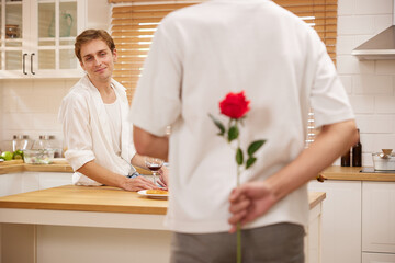 Fototapeta premium young gay couple looking at boyfriend and holding rose behind his back for surprise in the kitchen