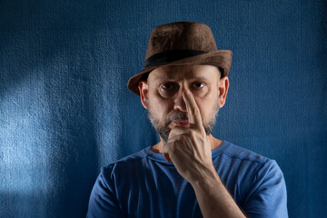 Studio portrait of man wearing hat and finger to nose looking at camera.