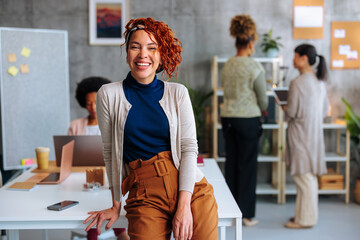 Portrait of young Hispanic business woman sitting on company office desk with coworkers in background. - 618300667