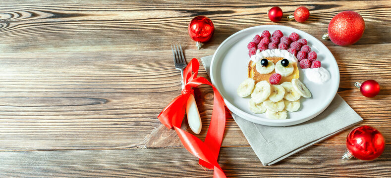Christmas Santa Claus face shaped pancake with sweet fresh raspberry berry and banana on plate wooden background for kids children breakfast dinner. xmas food dessert new year decorations, red balls