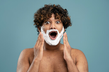 Excited Indian man with shaving foam on face isolated on blue background, looking at camera