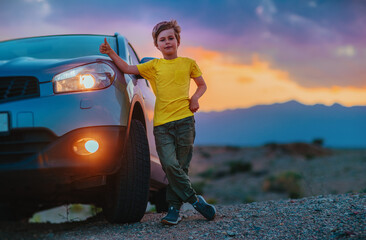 Naklejka premium Successful boy standing by the car and showing thumbs up at twilight on mountains background