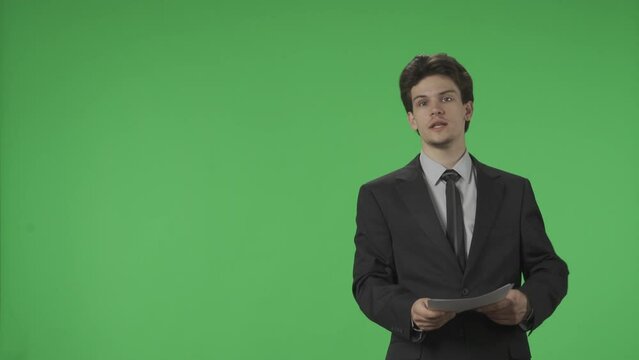 Reporting TV presenter with papers in his hands in a studio against a green screen. Anchorman speaks and points to the green screen. Advertising area, workspace mock up. HDR BT2020 HLG Material.