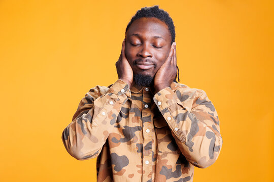 Man with close eyes covering ears with palms, doing three wise monkeys gesture in front of camera over yellow background. African american person not listening to noise and not speaking in studio