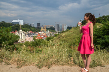 Full length portrait of young beautiful dark-haired woman wearing pink dress holding a bottle of water in hand and enjoying the city view