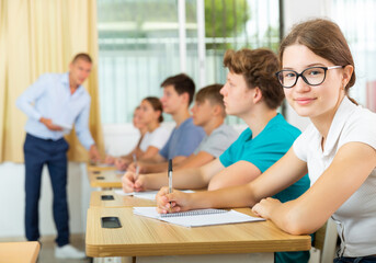 Cute smiling teenage schoolgirl in glasses sitting at table in classroom during lesson, writing lectures in workbook