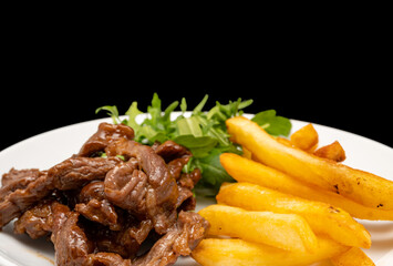 Stir-fried beef served with french fries and arugula leaves on white plate, isolated on black background. Close-up view.