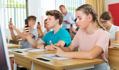 Teenager students sitting in class room with smartphones and photographing something.