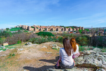 Fototapeta na wymiar Ciudad medieval de Ávila con dos turistas de espaldas sin identificar mirándola, un día soleado con cielo azul, desde Castilla y León , España.