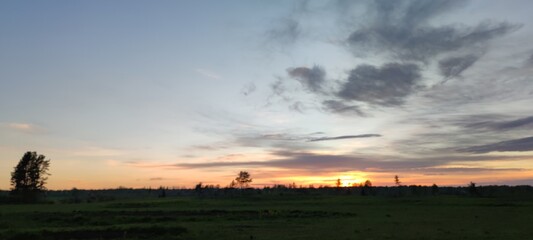 Spring sunset over the field. The sun has gone below the horizon, its last rays are visible. There are high gray clouds in the blue sky. Below them is a dark field and the outlines of growing trees.