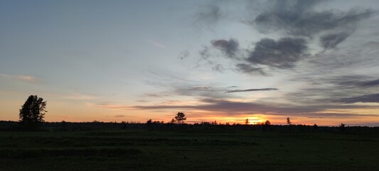 Spring sunset over the field. The sun has gone below the horizon, its last rays are visible. There are high gray clouds in the blue sky. Below them is a dark field and the outlines of growing trees.