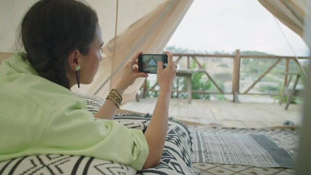Young woman lying in comfortable glamping tent and taking photos of nature with mobile phone