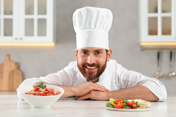 Happy professional chef near delicious food at marble table in kitchen