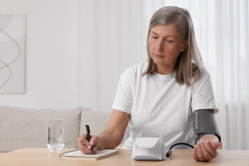 Woman measuring blood pressure and writing it down into notebook at wooden table in room, space for...