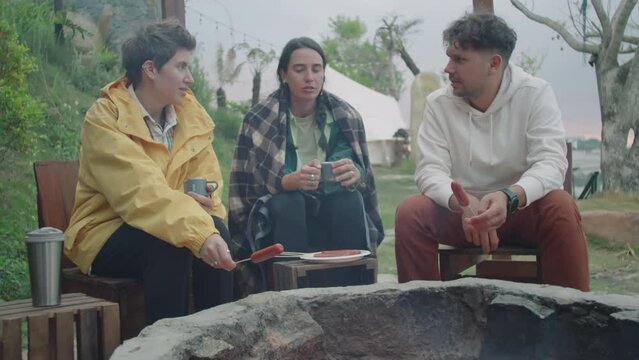 Medium full shot of company of young tourists cooking sausages over campfire flame and having conversation while glamping in nature