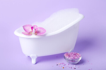 Fototapeta na wymiar Small bathtub with foam, sea salt and orchid flower on lilac background