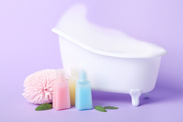 Fototapeta na wymiar Small bathtub with foam, sponge and bottles of cosmetic products on lilac background
