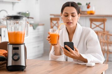 Young woman with glass of vegetable juice using mobile phone in kitchen