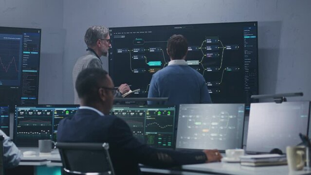 Multiracial team of IT technical specialists and software engineers work with data server, live analysis charts and blockchain network on computers and big digital screens in modern monitoring office.
