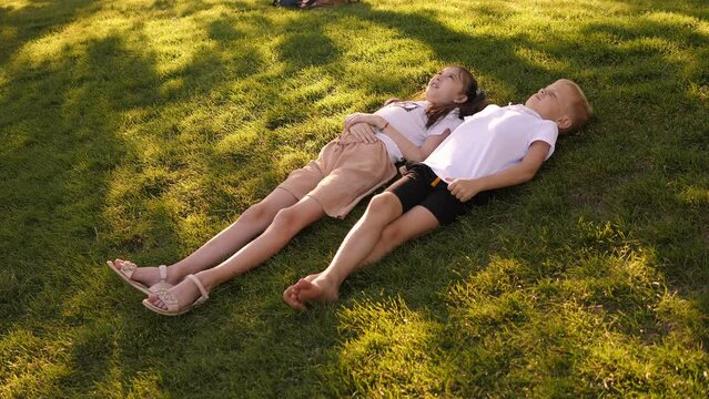 Cheerful boy and girl lie in the park on the green grass in summer and talk. Children are relaxing in the park at sunset.