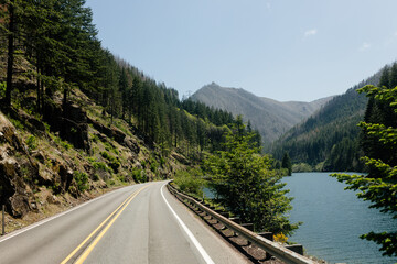 A beautiful road in the mountains among trees, on the right is a large lake with blue water. Summer landscape with an asphalt highway, mountains, trees and a river. Nature in Oregon in spring.