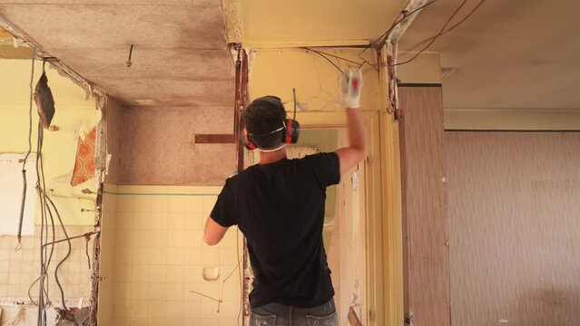 Man breaking wall and door frame inside demolition apartment with sledge hammer, static wide shot