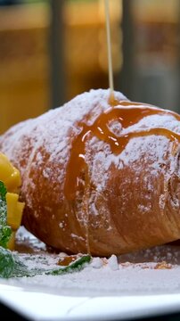 watering croissant with liquid caramel on white plate mango mint leaves and delicious french dessert breakfast lunch thin stream of sweet syrup pouring on bun pastries fresh food on hotel balcony