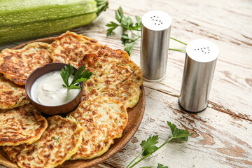 Plate of tasty zucchini fritters with sour cream on light wooden background