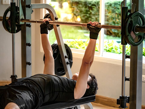 Active Asian sport man in black sportswear lying on exercise bench doing barbell bench press lifting at weightlifting station. Indoor weight training and muscular building workout in fitness gym