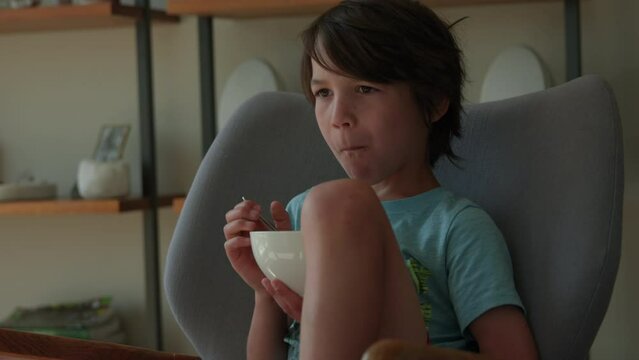Young boy eating ceral while sitting in living room chair looking off towards tv - medium shot