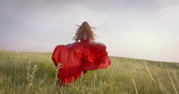 Happy joyful beautiful young woman in medieval vintage dress is runing in field, enjoying nature. Slow motion. Red vintage dress. Long red hair fly flutter in wind. Cheerful woman runs, back rear view