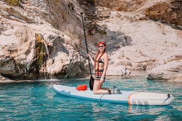 Happy girl in a swimsuit with a paddle swims on a SUP board on the sea near huge limestone rocky cliff. Healthy lifestyle and recreation concept