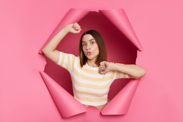 Beautiful caucasian brunette woman wearing striped t shirt posing in torn pink paper wall standing...