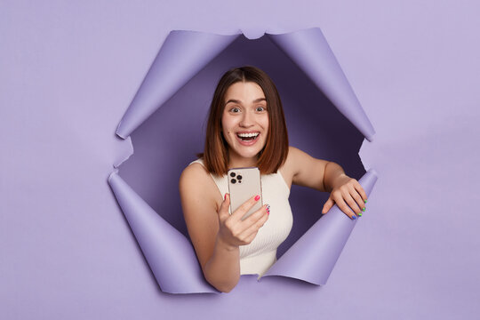 Extremely happy joyful cheerful young brunette woman in casual clothing breaking through purple paper hole using mobile phoine checking social networks.
