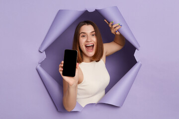 Excited cheerful young brunette woman in casual clothing breaking through purple paper hole showing cell phone with empty screen copy space for advertisement.