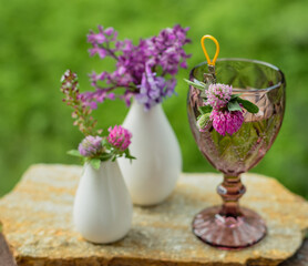 A glass with a drink and flowers in a white vase on the terrace against the backdrop of a green garden