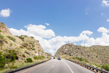 Fototapeta premium Beautiful highway with cars and trucks among mountains on a sunny day with blue sky and fluffy clouds. A summer landscape with the road among mountains with rocks and grass