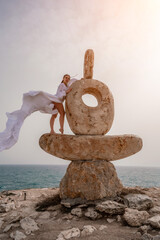 Fototapeta na wymiar Woman sea stone. A woman stands on a stone sculpture made of large stones. She is dressed in a white long dress, against the backdrop of the sea and sky. The dress develops in the wind.