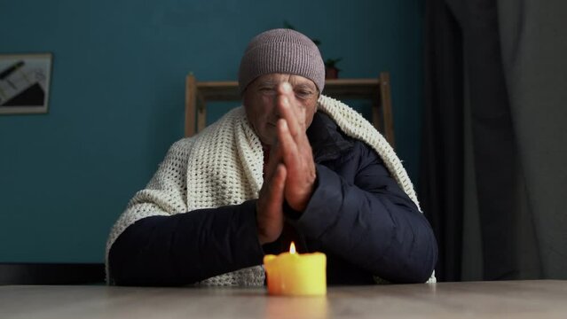 Senior man dressed in a warm winter hat and a scarf sits at home at a table and warms his hands from a burning candle. Concept of no heating in winter at home.