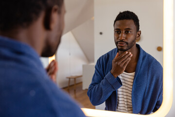 Happy african american man in bathrobe looking in mirror in bathroom