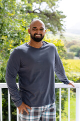 Portrait of happy biracial man in pyjamas standing on sunny balcony at home in front of treetops