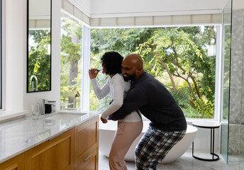 Happy biracial couple in pyjamas having fun embracing and brushing teeth in bathroom in the morning
