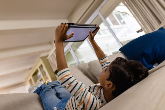 Low angle of happy biracial boy lying on couch using tablet with copy space on screen in living room